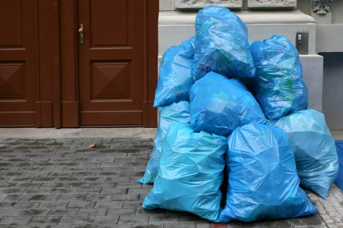 A group of several large blue plastic rubbish bags filled with waste materials are stacked on a paved pavement surface in front of a residential property. The bags are gathered near a brown wooden door and a grey stone wall, with some of the bags leaning against each other and others positioned on the ground. The bags have a slightly crumpled texture, and their synthetic plastic material reflects natural daylight, suggesting an outdoor setting. The area appears to be a residential driveway or front pathway, where rubbish removal services, such as those offered by House Clearance Norbiton, might collect disposed waste. The scene indicates a typical rubbish collection point in Norbiton, with environmental context limited to the immediate pavement and building frontage, maintaining a neutral and professional description aligned with local rubbish management and clearance activities.